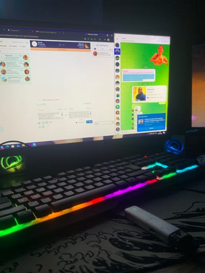 The image shows a dimly lit room with a computer setup. A desktop monitor displays a messaging application that appears to be a cross between a dating app and a social media platform. On the left side of the screen, a list of contacts is visible, with names like "Patrick," "Yyyyjohn," and "Jeff." The right side of the monitor shows a chat window with a user named "Jamal" and another conversation that includes text in Russian and English, suggesting a translation feature is in use. A keyboard with RGB lighting is positioned in the foreground, and a vape pen rests on a mousepad with a stylized design. The overall impression is of a personal workspace, likely in a home environment. The image does not provide any specific visual cues or context that would allow for the determination of its location within the city of Glodeni, Moldova.