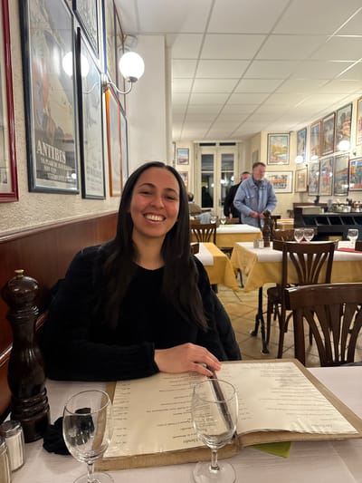 A person is sitting at a table in a restaurant. A menu is open on the table, displaying a list of dishes in French, alongside wine glasses and a pepper grinder. A woman with dark hair is smiling. Other people are seated at tables in the background. The restaurant's interior includes framed posters on the wall and light fixtures. There is no indication that this picture was taken in Chêne-Bourg, Switzerland.