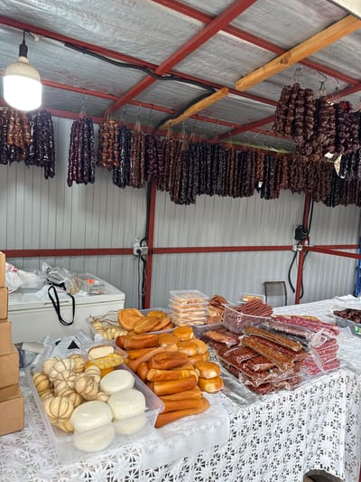 This is a medium shot of a food stall, likely at a market, showcasing a variety of cured meats and sweets. The stall is set up indoors, with a corrugated metal wall in the background and a red metal frame structure supporting the ceiling. To the left, numerous strings of dark, glistening treats, possibly dried fruits or candies like churchkhela, hang from the ceiling. These appear to be stacked in dense rows, varying in color from deep brown to black.

In the center and right of the image, the stall features a display of packaged goods. There are several clear plastic containers holding various types of cheese, some resembling small balls tied with twine, and others appearing as larger, round loaves. Adjacent to the cheese are rows of bread-like items, possibly baguettes or rolls, and several packages of smoked sausages of different sizes and colors, some appearing to be whole links and others sliced. A white, intricately patterned tablecloth or fabric is visible beneath some of the products on the right.

A bright LED light fixture is attached to the metal frame on the left side, illuminating the scene. The overall impression is one of abundance and variety, suggesting a bustling food market environment. There are no people visible in the frame, and the lighting suggests it could be daytime, though the indoor setting makes it difficult to determine specific weather conditions. No discernible text is visible on the products or stall in the image. The location context provided is Guamka, Russia.
