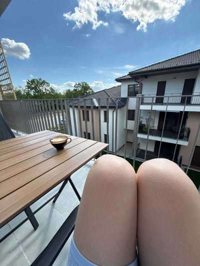 A view from a balcony, oriented sideways, shows the lower bodies (thighs) of a person sitting, a wooden table, and a cityscape beyond the balcony railing. The thighs, filling the right side of the frame, are bare and appear Caucasian in skin tone. To the left, a table with a light brown wooden surface is visible. On the table, there is a dark-colored cup containing a light liquid. The balcony railing is made of metal bars. Beyond the railing, buildings are visible under a blue sky dotted with white clouds. Trees are interspersed among the buildings. Based on the architectural style and surroundings, it is potentially located in a residential area within Balatonlelle, Hungary.
