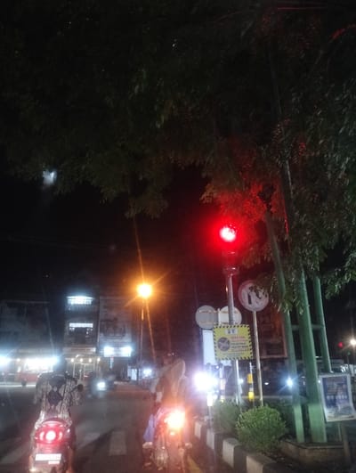 The image depicts a nighttime street scene in Gampa, Indonesia. The primary subjects are individuals riding motorcycles. The setting is an urban street, evident from the buildings and streetlights. A prominent red traffic light is illuminated, indicating a stop. There are also road signs visible, one of which is a yellow sign with black text that appears to be an announcement or regulation. The time of day is clearly night, with artificial lights from buildings and vehicles illuminating the scene. The weather is not explicitly indicated, but the darkness suggests it's not daytime. The overall mood is functional, depicting the common activity of nighttime travel in an urban environment. The presence of multiple motorcycles and pedestrians suggests a relatively active street, even at night. One building has a bright white light emanating from its upper floors, and another has illuminated signage. The trees in the foreground are silhouetted against the night sky and the artificial lights.
