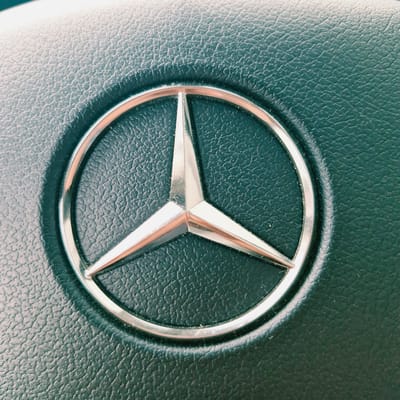 The image shows a close-up of a Mercedes-Benz logo on a dark, textured surface, likely leather from a steering wheel or dashboard. The logo is a silver three-pointed star enclosed within a circle. The background exhibits a fine, irregular texture. There are no people visible in the image. The lighting creates subtle shadows around the logo, highlighting its metallic sheen and the texture of the surrounding material. The location of this object within the city of Başakşehir, Turkey, cannot be determined from the image itself.