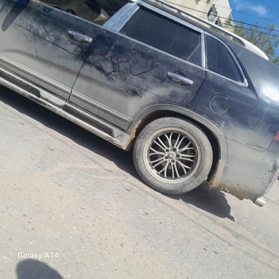 A black, dirty SUV is parked on an asphalt street in what appears to be Nuseirat, Palestinian Territory. The vehicle has roof rails and side steps. The rear tire is visible with a multi-spoke rim. The car's body is covered in dirt and scratches. The surrounding area is a paved street with a building and some trees visible in the background.