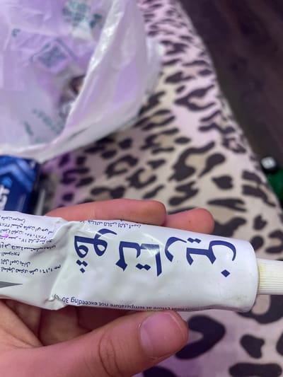 The image shows a close-up of a white tube of ointment with Arabic text and some English text. The tube is held by a person's hand, with fingers visible on the right side. The ointment tube has blue Arabic lettering that appears to be a brand name, and also some smaller text in Arabic and English. Part of a white plastic bag is visible in the background, as is a patterned fabric. The English text on the tube includes "Store not exceeding 30 C". There is no information or visual evidence in the image that indicates the location is Vaganovo, Russia.