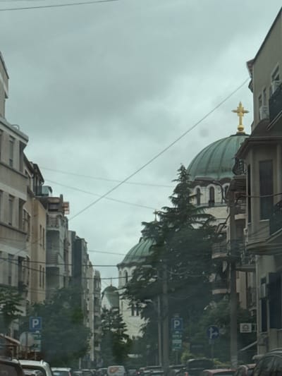 The image depicts a view down a city street with buildings on either side and a large domed church in the background. The sky is overcast and gray. The buildings appear to be residential or commercial, with multiple stories and windows. There are trees lining the street and some power lines are visible overhead. In the foreground, several cars are present, suggesting that the photo was taken from a moving vehicle. There are also street signs visible, including one with a blue "P" indicating parking. The architecture of the domed church, particularly its green dome and golden cross, is distinctive and characteristic of Orthodox Christian churches. The overall impression is of an urban environment with a significant religious structure at its center. The scene is consistent with Vracar Urban Municipality in Serbia, known for its churches and residential buildings.