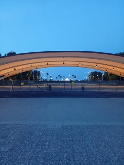 An exterior, eye-level, medium shot captures an arched canopy structure in Civitanova Marche, Italy, at dusk. The canopy, made of a light-colored fabric stretched over a curved frame, is illuminated from beneath by rows of hanging black lights, creating a warm glow. The sky above is a deep, clear blue, transitioning to a lighter hue near the horizon.
Beyond the canopy, a fence made of metal posts and wire mesh encloses an area. Behind the fence, a line of palm trees stands silhouetted against the twilight sky, interspersed with streetlights. The ground in front of the canopy is paved with large, rectangular concrete slabs, while the area further back appears to be asphalt. There are no people or animals visible in the scene. The overall impression is one of an empty, transitional space at the edge of a developed area.
