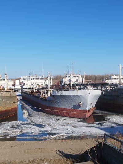 The image shows several ships docked in a partially frozen body of water, likely a river or canal. The largest ship in the foreground is predominantly gray with a red waterline stripe. The water contains ice floes and reflections of the vessels. The foreground also includes a sandy bank with sparse vegetation. The scene suggests a cold climate or season. The ships are likely in Zhatay, Russia based on the prompt.