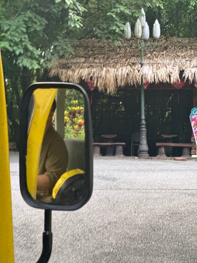 The image shows a side-view mirror of a vehicle, reflecting a person wearing a light brown shirt. The mirror is framed in black plastic and is mounted on a black arm. To the left of the mirror is a bright yellow object, possibly part of the vehicle. In the background, visible through the mirror and to the right, is a thatched-roof structure with a lamp post in front of it. The structure appears to be an open-air pavilion or shelter with stone benches. Red decorative elements hang from the roof. Beyond the structure, lush green foliage suggests a natural or park-like environment. The ground in the foreground is paved with asphalt. The overall impression is that the photograph was taken from within a vehicle moving through a scenic area, possibly a tourist attraction in Tây Hoa Lư Ward, Vietnam, given the architectural style of the shelter.