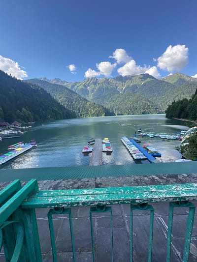 The image shows a wide view of Lake Ritsa in Abkhazia, Georgia. In the foreground, a weathered green metal railing with peeling paint overlooks a green corrugated roof. Beyond the roof, the turquoise water of the lake is visible, with several docks filled with colorful paddle boats. Some of these boats are yellow, red, green, and blue, and many are unoccupied. A few people are visible on the docks and one person is on a paddle boat. The lake is surrounded by steep, tree-covered mountains, with some of the higher peaks showing patches of snow. The sky is a bright blue with scattered white clouds. A few small boats and some buildings are visible on the far shore of the lake. The image captures a scenic natural landscape with recreational activities.