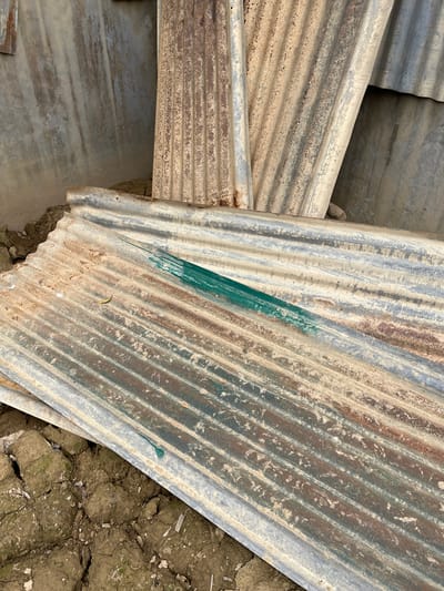 The image shows several sheets of corrugated metal, some of which appear weathered and rusty, piled on the ground. The ground is composed of dry, cracked earth with some patches of moss or dirt. In the background, a concrete structure with an uneven, textured surface is visible. One sheet of corrugated metal in the foreground has a distinct diagonal streak of green paint. Another sheet has a small, yellow leaf lying on it. The overall scene suggests an outdoor area, possibly a construction site or storage area, in Benuaraja, Indonesia, with the materials suggesting a utilitarian or possibly neglected setting.