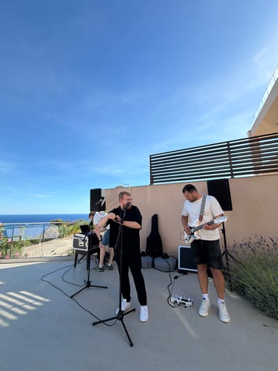 A band is performing on a rooftop terrace in Krimovice, Montenegro, with a clear blue sky overhead. Two men are the primary subjects. The man on the right is wearing a white t-shirt and denim shorts and playing a white electric guitar. He is looking down at the guitar as he plays. The man on the left is wearing a black shirt and black pants, and is singing into a microphone. He has glasses and a beard. A third person, partially obscured, is behind a keyboard and mixing desk, also appearing to be part of the band. In the background, a body of water, likely the Adriatic Sea, is visible beyond a railing and some greenery. The terrace itself is paved with light-colored tiles. There are speakers, microphones, and music equipment set up for the performance. A guitar case and a few other bags are visible on the ground behind the musicians. A modern building with a balcony railing is visible to the right of the terrace. A dense patch of lavender is growing to the far right of the frame. The overall scene suggests an outdoor musical performance on a sunny day.