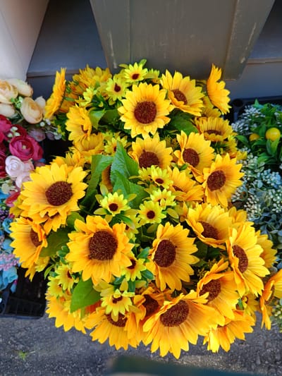 The image shows a close-up, overhead view of a large bouquet of artificial sunflowers and other yellow flowers. The sunflowers are the most prominent, with their bright yellow petals and dark brown centers. They are arranged in a dense cluster, with some smaller yellow flowers interspersed among them. To the left of the sunflowers, there are some artificial roses in shades of pink and red, along with some light pink and cream-colored flowers. In the background, to the right, there is a display of greenery with small white flowers and some artificial yellow fruits, possibly lemons. The flowers appear to be arranged in a planter or bin, with the edge of a dark container visible at the bottom left. The ground beneath the flowers appears to be asphalt or gravel. The scene is well-lit, suggesting it is outdoors or in a brightly lit space. There are no people or animals visible in the image. The overall impression is one of a colorful and vibrant floral display, likely for sale or decoration.