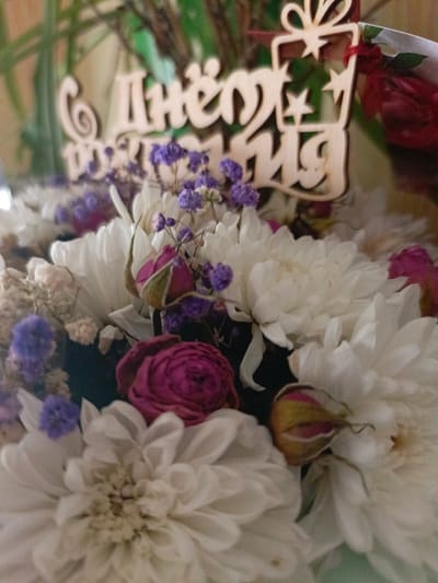 The image shows a close-up of a bouquet of flowers with a wooden decoration. The bouquet contains white chrysanthemums, pink roses, and small purple flowers. The wooden decoration is in the shape of a gift box with stars and appears to have text on it, though it is partially obscured. The lighting suggests an indoor setting. There is no indication of a specific location in the city of Zalesovo, Russia.
