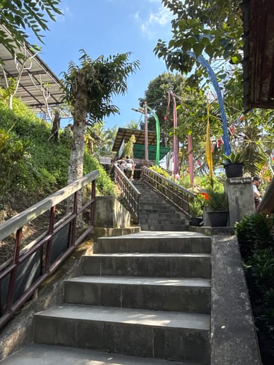 In Tampaksiring, Indonesia, a stone staircase ascends a lush, verdant hillside under a bright blue sky. The stairs are bordered by a sturdy wooden and metal railing on the left and a concrete embankment on the right, which is adorned with potted plants. At the top of the stairs, a building with a green roof is visible, and colorful flags, including yellow, blue, and pink, flutter in the breeze, adding a festive atmosphere to the scene. Several individuals are seen walking up the stairs and gathered near the building. The area appears to be a tourist attraction or a place of worship, with its natural beauty enhanced by decorative elements.