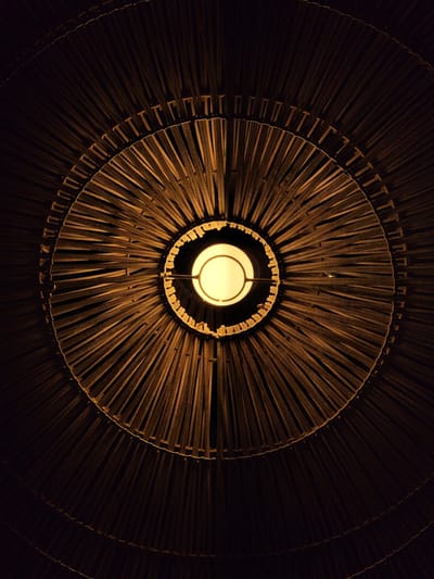 This image shows a close-up, overhead view of a light fixture. The fixture is round and appears to be made of natural materials, possibly wood or reeds, arranged in a radial pattern. The material is dark brown and creates a textured surface with distinct shadows. Concentric rings are visible within the structure, adding depth and complexity. At the center of the fixture, a bright, round light bulb is illuminated, casting a warm glow and creating dramatic highlights and shadows on the surrounding material. The overall impression is one of abstract beauty and the interplay of light and shadow. The setting appears to be indoors, given the artificial light source and the absence of any environmental context suggesting an outdoor location. There is no text visible in the image.