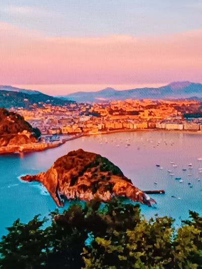 The image depicts a panoramic view of a coastal city at sunset. In the foreground, lush green foliage partially obscures the view of a body of water. The water is a vibrant blue and is dotted with numerous small boats.

Dominating the middle ground is a prominent, rocky island covered in greenery and brown vegetation, jutting out into the water. A dark structure, possibly a pier or a small building, extends from the island.

Beyond the water lies the city, characterized by a dense collection of buildings with orange-tiled roofs. The cityscape stretches along the coastline, nestled against a backdrop of rolling hills and mountains in the distance.

The sky is painted with soft hues of pink and orange, transitioning to a pale blue at the top, indicative of a sunset or sunrise. The overall impression is one of a picturesque coastal landscape bathed in the warm glow of twilight. This scene is likely a view of La Concha Bay in San Sebastián, not Vitoria-Gasteiz.