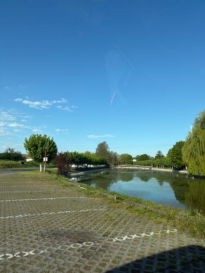 A low-angle landscape oriented view of a canal scene with a blue sky and clouds, taken in Târgu Mureș, Romania. The canal runs horizontally across the top right quadrant. Reflections of trees and the sky are visible in the water. The near side of the canal is bordered by grass and a grey block paved area, with white painted parking space markers. A red and white barrier and some trees are visible in the lower center of the image. The sky occupies a large area in the left side of the image. The view is from inside a vehicle, with some internal reflections visible.