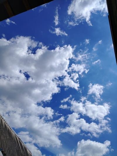 The image depicts a view looking upwards at a bright blue sky filled with white cumulus clouds. A portion of a gray, corrugated roof structure is visible in the bottom left corner, angled upwards and to the left. The upper right corner of the image shows a dark, vertical element, possibly a building edge or a frame. The lighting suggests it is daytime, with sunlight illuminating the clouds. The scene is an outdoor environment, likely a rooftop or an open area with a view of the sky. There are no people or discernible actions taking place. The image is a photograph taken from a low angle, emphasizing the vastness of the sky. There is no information available to specifically place this image in the city of Lipetsk, Russia.