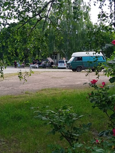 The image shows an outdoor scene in Luka, Ukraine. In the foreground, lush green grass and rose bushes with red blossoms are visible. Further back, a gravelly area is present with several people standing around tables. A dark-colored sedan and a blue and white van are parked in the vicinity. Behind these, a building with windows and a sloped roof can be seen. Tall trees with green foliage dominate the background, with some branches and leaves extending into the foreground. The sky is overcast.