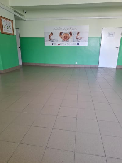 The image depicts an interior hallway with green and light-colored walls. On the left wall, a framed poster with anatomical diagrams is mounted. In the center, a large banner titled "Akademia Rodzica w Zabrzu" (Parent Academy in Zabrze) is displayed, featuring images of pregnant women and babies. To the right, a white door is visible, with a sign indicating "POMIESZCZENIE GOSPODARCZE 64" (Utility Room 64). The floor is tiled with light brown squares. A security camera is mounted on the ceiling. The environment appears to be a public or institutional building, likely within Zabrze, Poland, given the banner's text. There are no people visible in the image.