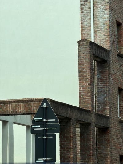 The image shows the rear of a building constructed with red brick and off-white stucco. A dark triangular sign with white text and a metal pole holding multiple dark rectangular signs is visible in the foreground. The signs appear to be traffic or informational signs. The building has architectural details including brick columns, window openings, and a sloping roofline. The scene is an exterior urban environment, possibly a side street or alleyway, given the focus on the building's rear and the presence of signage. The lighting suggests it is daytime. The image does not contain any people or observable actions. The location in Évry-Courcouronnes, France is not visually evident from the image itself, but could be inferred from external information not present in the visual data.