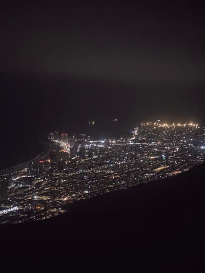 This is a nighttime aerial view of a coastal city, likely Alto Hospicio, Chile, with the Pacific Ocean on the left. The city is densely populated, with numerous buildings illuminated by lights. The lights create a sparkling effect across the urban landscape, highlighting streets and structures. A beach runs along the coastline, with waves visible as they approach the shore. In the distance, a few ships or boats are visible on the ocean, their lights also twinkling. The overall impression is of a vibrant, active city at night.