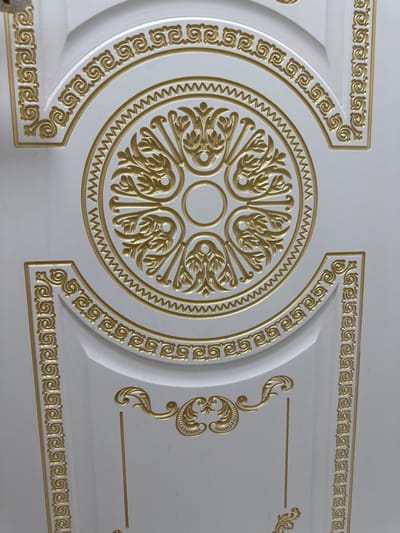 The image shows a close-up, overhead view of a white, textured ceiling with ornate gold embellishments. The dominant feature is a large circular medallion in the center, composed of intricate gold scrollwork and leaf-like patterns. This medallion is surrounded by a toothed border, and further out, by another gold border with a repeating scroll motif. Below the central medallion, a smaller, more delicate gold design with curved lines and leaf shapes is visible. The ceiling itself appears to be made of panels, with visible seams and a subtle, undulating surface. The lighting is even, highlighting the three-dimensional nature of the gold detailing against the matte white background. There are no people or other objects present in the frame. This decorative ceiling element is located in Dushanbe, Tajikistan, likely within a building of significant architectural or historical importance, such as a government building, cultural institution, or a high-end residential or commercial space.