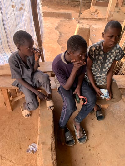 Three male juveniles are observed seated on a wooden bench within an outdoor or semi-sheltered area in Ube, Nigeria. The ground consists of uneven, reddish-brown earth.
The juvenile on the left wears a dark grey long-sleeved shirt and trousers with beige slide sandals, his right hand positioned near his mouth. The central juvenile wears a dark purple t-shirt and dark trousers with side stripes. He is barefoot, with dark slide sandals on the ground nearby, his right hand covering his mouth, and his left hand holding a green device with a red elastic band. The juvenile on the right wears a grey and dark checkered short-sleeved shirt and light brown trousers. He is barefoot, with dark slide sandals on the ground nearby, and holds a stack of Nigerian Naira banknotes in his left hand.
In the background, a light-colored striped fabric screen is visible on the left, and eroded earthen structures or steps are present towards the right. A small, translucent plastic bag containing unidentifiable contents rests on the ground in the foreground.