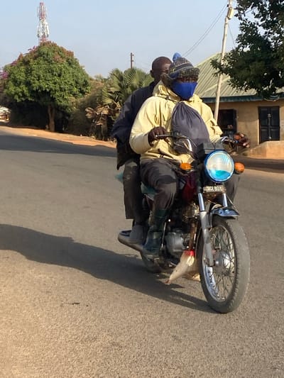 Two adult males are riding a red motorcycle on an asphalt road in Udu, Nigeria, under direct sunlight, casting long shadows to the left. The driver, wearing a light yellow jacket, dark trousers, dark green rubber boots, a blue face mask, and a knitted beanie with a blue pom-pom, grips the handlebars. A dark blue drawstring bag hangs from his neck. The passenger, wearing a dark jacket and trousers, sits behind the driver. The motorcycle has a round headlight, partially obscured by a light blue plastic covering, with "NICE BOY" inscribed below it, and orange turn signals. A white plastic bag is tied near the front wheel. In the background to the left, a large green tree with some red foliage stands, with a white cellular tower visible above it. To the right, additional trees and a light-colored building with a corrugated metal roof are present.