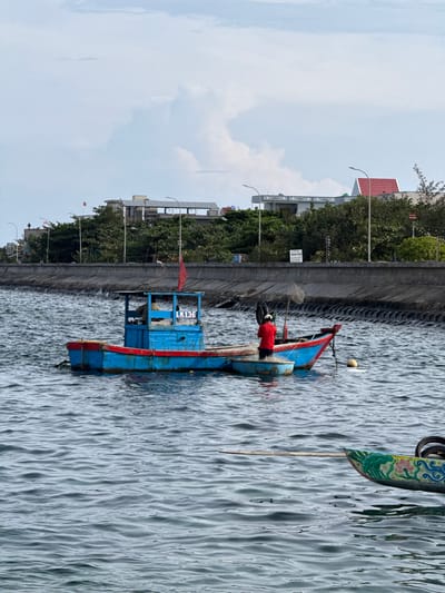 A blue and red fishing boat with a covered cabin is anchored in choppy, grey water. A person wearing a red shirt and white helmet stands on a smaller, attached boat, holding a fishing net. The boat is labeled "LK136". In the background, a concrete seawall runs along the shore, topped with trees and buildings. A red flag flies from the cabin of the fishing boat. The sky is overcast with scattered white clouds. Another decorated boat with a green dragon design is partially visible in the foreground.