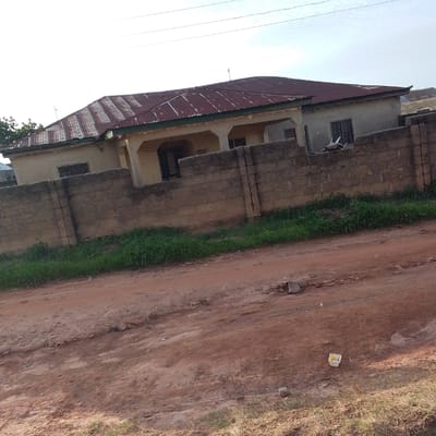 This media file depicts a residential scene in Mangu, Nigeria. The central focus is a single-story house with a corrugated metal roof, appearing weathered and aged. The house has a light-colored exterior and is partially obscured by a low, concrete block wall that runs along the front of the property. The wall is made of rough, stacked blocks, some of which show signs of wear and discoloration.
In front of the wall, a patch of overgrown, green grass is visible. Beyond the wall and the grass, a dirt road or pathway dominates the foreground. This road is reddish-brown in color and appears uneven, with scattered stones and debris. A white plastic container, possibly a bucket or bin, lies on the road near the grass.
The sky above is overcast, suggesting a cloudy or hazy day. The lighting indicates it is likely daytime, possibly late afternoon or early morning due to the diffused light. There are no people visible in the scene, and the overall impression is one of a quiet, rural or semi-urban setting. The scene conveys a sense of everyday life rather than a specific event. There is no visible text within the image.