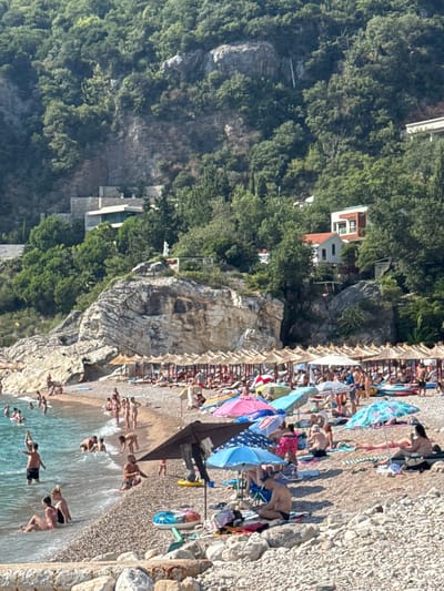 The image captures a lively scene at a pebbly beach in Perazića Do, Montenegro, bathed in bright, sunny daylight, suggesting a warm, pleasant day. The beach is densely populated with people enjoying various summer activities. Many are lounging on beach chairs or towels, some under colorful umbrellas, indicating a relaxed atmosphere. A significant number of people are in the water, some wading and others diving or swimming. The clear, turquoise water is inviting, with gentle waves lapping the shore.
In the background, lush green trees and rocky cliffs rise steeply, adding a dramatic natural backdrop to the beach scene. A few buildings are visible nestled amongst the trees on the hillside, hinting at the proximity of residential or tourist facilities. The overall impression is one of a popular holiday destination, buzzing with activity and the joy of summer leisure.