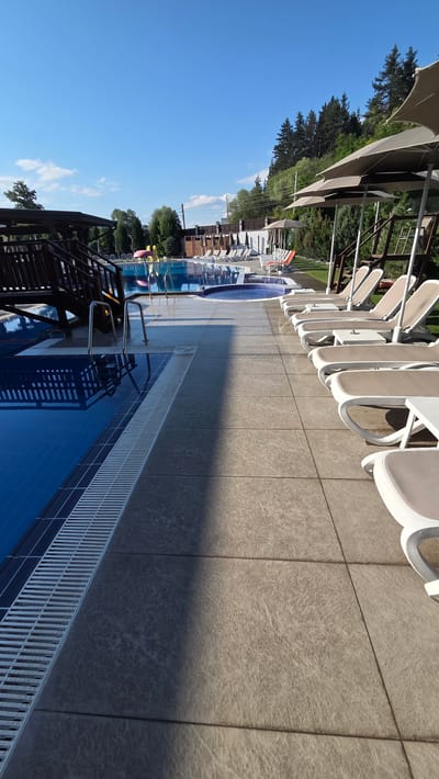 The image shows an outdoor swimming pool area with lounge chairs and umbrellas under a clear blue sky. The pool is blue and appears to be empty. There is a wooden structure with stairs next to the pool. In the background, there are trees and a fence. The area is paved with large, light-colored tiles. The image is a photograph of a resort in the city of Bania, Bulgaria.