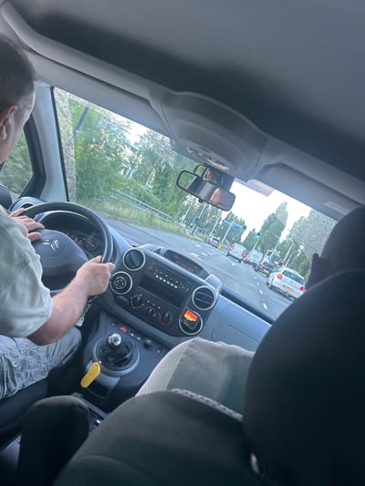 The image is taken from inside a vehicle, looking forward through the windshield at a street scene in Rijswijk, Netherlands. A person's hands are visible on the steering wheel of the car, indicating they are driving. The dashboard of the car is visible, with a gear shift and various controls. In the background, other vehicles are on the road, and traffic lights are visible. Trees and buildings line the street. The weather appears to be overcast, with some water droplets on the windshield.