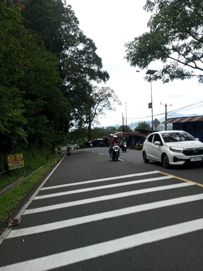 The scene depicts a street view within the city of Phoxay, Laos, under an overcast sky. In the foreground, a dark asphalt road features a prominent pedestrian crosswalk with four thick white stripes. A faint yellow line runs parallel to the right edge of the crosswalk. On the left side of the road, dense green foliage covers a steep embankment, and a yellow sign with black text, partially obscured, reads "HATI - HATI JALAN BERLUBANG" (Indonesian for "BE CAREFUL - POTHOLED ROAD").

Traffic is present on the road. A white Honda Brio compact hatchback, bearing a license plate that appears to read "B 3508 ZOU," travels on the right lane, moving towards the viewer's right. Ahead of the car, a dark motorcycle carrying two individuals, both wearing helmets (the driver in a red shirt, the passenger in a maroon shirt), proceeds away from the viewer in the middle lane. Several other motorcycles are visible further down the winding road, also moving away from the viewer, with at least one making a turn to the right.

On the right side of the road, behind the white car, a utility pole with a solar panel fixture is visible, extending above trees. Further in the background, more utility poles with overhead wires are present, alongside various small structures with corrugated metal roofs, suggesting commercial or residential buildings. Distant hills or mountains are faintly discernible on the horizon. The road surface appears damp in areas.