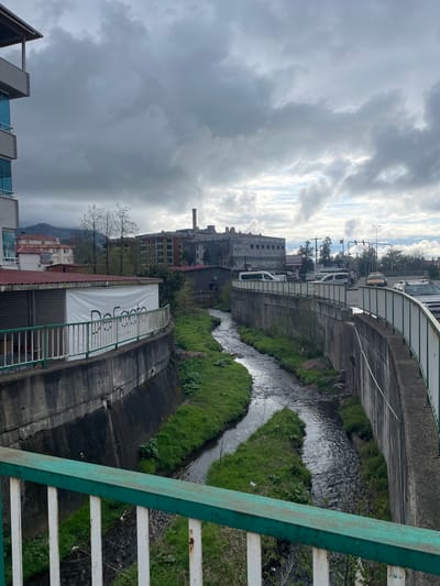 The image depicts a creek running through a channel in Kemalpaşa, Turkey. The water flows from the upper right towards the bottom left of the frame. The channel is lined with concrete walls and vegetation, including grass and some shrubs. Buildings line the channel on both sides, with some appearing residential and others possibly commercial or industrial. A bridge crosses over the creek in the upper right of the frame, with traffic visible on the road above. The sky is overcast with heavy clouds. Part of a green and white painted railing is visible in the lower and right foreground. A sign with "De Facto" written on it is visible on a building next to the creek. The photo appears to be taken from an elevated position on a bridge or higher structure.