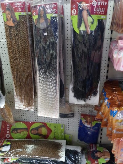 The image depicts a shelf displaying various synthetic hair extensions. Packaged hair extensions, including a brown "Twist 'N Curl" and a black-to-white ombre style, hang from hooks. Brands like "Aviv" and "Lulu Curls" are visible on the packaging. Below, more extensions are stacked, including a green pack labeled "Diva Twist." Cosmetic products, possibly skincare, are seen on the right side of the shelf. The display has a diverse selection of hair textures and colors.