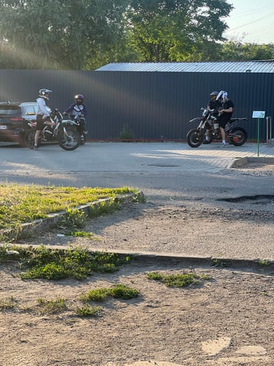 Four individuals, two on motorcycles and two standing near motorcycles, are gathered in an outdoor paved area. The individuals are wearing helmets, suggesting they are motorcyclists. One motorcycle, a white and orange dirt bike, is positioned to the left of the frame, with two people interacting beside it. A dark-colored SUV is parked behind this motorcycle. To the right, another motorcycle, black and orange, is occupied by two individuals. A dark, corrugated metal wall forms the backdrop for the scene, with a metal roof structure above it. Trees with green foliage are visible in the upper portion of the image. The ground in the foreground consists of dirt and patchy green vegetation. The overall scene appears to be a casual gathering of motorcyclists. This specific scene is not identifiable as being in Kropyvnytskyi, Ukraine.