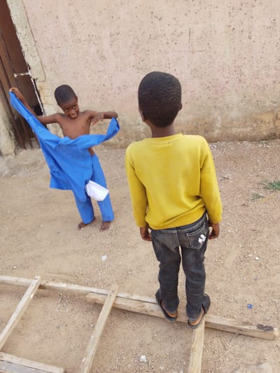 Two young boys stand in a sandy, dusty area next to a pale wall. One boy, shirtless, stands holding a blue outfit with a white cap attached. He is smiling and appears to be putting the outfit on. The second boy is standing facing away from the camera, wearing a yellow long-sleeved shirt, dark jeans, and sandals. He is standing on a wooden frame on the ground. The location appears to be outside, potentially in the city of Bauchi, Nigeria.
