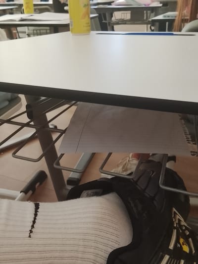 The image captures a first-person perspective from under a desk in what appears to be a classroom or study environment in Petrich, Bulgaria.

In the immediate foreground, a person's lower leg and foot are visible. The leg is covered by a white, ribbed, elastic-looking material, possibly a medical brace or a thick sports sock. Attached to the foot is a black and white sports shoe or skate, featuring intricate white lacing or straps, black plastic components, and some yellow details/logos.

Above the foot, the underside of a white tabletop with a dark edge is prominent. Directly below the table, a metal frame, likely part of the table's support structure, is visible. A piece of lined paper, possibly from a notebook, hangs or rests on this frame.

On the surface of the white tabletop, a yellow bottle or can, partially obscured, is visible towards the right. Faint text "BLACK TEA" is discernible on its label. Another thin, blue object, possibly a pen or ruler, lies flat on the table's surface further left.

In the background, other tables and chairs, typical of a classroom setting, can be seen, suggesting multiple individuals in the space. The flooring appears to be a light-colored wood or laminate. The overall lighting suggests an indoor daytime setting. On the lined paper under the table, some faint, illegible writing can be seen along the right edge.