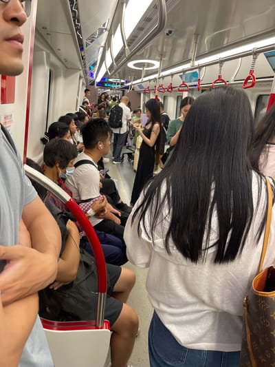 This media file shows the interior of a subway car, likely in Panyu District, China. The scene is crowded with people, with most individuals either sitting or standing. The perspective is from within the subway car, looking forward and towards the right.
The subjects in the image are primarily people. In the foreground, a person's face and arms are visible, looking upwards. Behind them, several individuals are seated. One person in the mid-ground, with long black hair, is wearing a white shirt and blue jeans, and has a Louis Vuitton-style bag on their shoulder. Other passengers are visible throughout the car, some looking at their phones, others talking, and some appearing to be resting. The subway car itself is characterized by its white interior, metal poles with red handles for passengers to hold onto, and bright fluorescent lighting overhead.
The activity taking place is the daily commute on public transportation. The subway car appears to be in motion, indicated by the blur of motion in some areas and the general busyness of passengers. There are no clear indicators of the time of day or the weather outside. However, the indoor lighting suggests it could be daytime or evening. The mood appears to be neutral, typical of a busy commute.
Notable details include the variety of clothing worn by passengers, from casual t-shirts and shorts to more formal attire. The presence of personal belongings like bags and phones is also evident. Some passengers are wearing masks, suggesting a potential ongoing health concern or personal preference. There is a small amount of visible text on a white sign near the bottom left of the image, but it is too small to be legible. The overall impression is that of a typical, crowded subway ride.