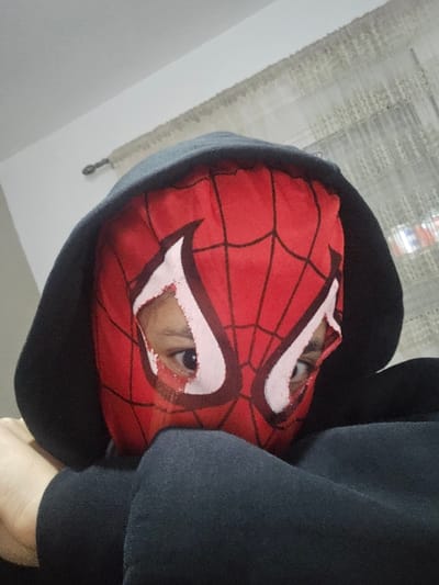A person wearing a Spiderman mask peers from a black hoodie. The mask is red with a web pattern and large, cartoonish white eye shapes outlined in dark red. The person's eyes are visible through the mask's eye holes. The hoodie's hood is pulled up, framing the mask. A portion of the person's arm and shoulder, clad in the black hoodie, is visible on the left. A sheer curtain hangs in the background.