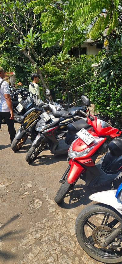 In an outdoor, sunlit scene in Bekasi, Indonesia, a row of parked motorcycles is visible on a paved surface. The motorcycles are predominantly scooters, with a mix of black and red models. One black Yamaha NMAX scooter is prominent in the foreground, displaying a license plate that reads "B 4171 KGZ." Another red Honda scooter is also visible, with a license plate reading "K 5465 KT." Two individuals are present. On the left, a person wearing a white t-shirt and dark pants stands facing away from the camera, their back to the viewer. To the right of the motorcycles, a young person with short dark hair, wearing a light green t-shirt, is looking to the right, away from the parked vehicles. The background features lush green foliage and trees, suggesting a location with ample vegetation. The paved ground appears somewhat worn and cracked.