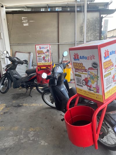The scene is located in Kecamatan Piyungan, Indonesia, at coordinates -7.824 latitude and 110.48 longitude. The image shows a street corner with two motorcycles parked. Both motorcycles are black and are in good condition. The motorcycle on the left is a Honda. The motorcycle on the right is attached to a red delivery cart. The delivery cart is emblazoned with advertisements for "Alfagift" and promotes "Bebas Ongkir." The cart has a red, cylindrical container, possibly for storing delivery items, located below the advertisements. Behind the motorcycles, a faded brick wall is visible, suggesting a possible alleyway or back entrance. The background appears to be a residential area with a building that seems to be a storefront, possibly a shop or restaurant.