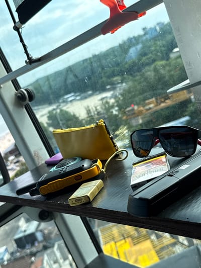A close-up, eye-level shot focuses on a wooden shelf placed near a window. The shelf displays several objects, including a walkie-talkie, a pack of Marlboro cigarettes, a Zippo lighter, and a pair of sunglasses. Behind these items, a yellow pouch and a purple object are partially visible. The window offers a view of a body of water, green trees, and buildings, suggesting an urban or suburban setting. The lighting is natural, coming from the window, and casts soft shadows on the shelf and objects. The overall impression is one of a relaxed, possibly solitary moment, with everyday items laid out casually. The presence of the objects and the outdoor scene visible through the window imply that this photograph was taken in Gdynia, Poland, likely during daylight hours.