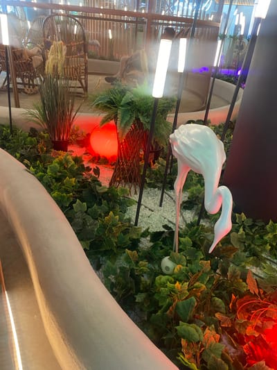The image depicts an indoor decorative scene featuring lush green artificial foliage, white gravel, and several illuminated cylindrical lamps. A large, white, abstract sculpture resembling a flamingo with a long, curved neck and legs is the central subject. The flamingo sculpture is positioned amongst the plants. In the background, wicker chairs are visible, suggesting an indoor seating area or lounge. A large, glowing red orb is also present, casting a warm light on the surrounding greenery. The lighting in the scene is a mix of cool white from the lamps and the warm red from the orb, creating a dramatic atmosphere. The overall impression is of a modern, artfully designed interior space. The image does not contain any elements that would allow for the specific location in Berdsk, Russia, to be determined.