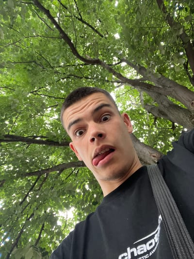 A close-up, low-angle selfie shows a young man sticking his tongue out and making a surprised face. He is wearing a black t-shirt with white lettering on the front and a grey strap is visible over his shoulder. He is positioned beneath a canopy of green leaves and tree branches, with sunlight filtering through the foliage. The background suggests an outdoor setting. There is no visible indication of the city of Sopot, Bulgaria.