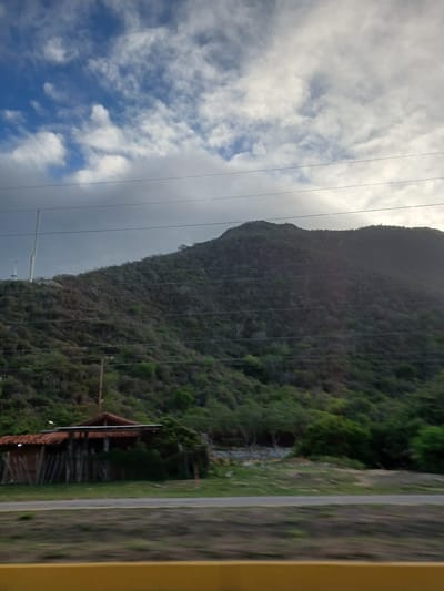 This image depicts a view from a moving vehicle in La Asuncion, Venezuela, showing a landscape dominated by a large, vegetated hill or mountain. The sky above is partly cloudy with scattered white clouds against a blue backdrop, transitioning to overcast conditions towards the right side of the frame. Several power lines stretch horizontally across the upper portion of the image, partially obscuring the sky and the hillside.
At the base of the hill, a small, rustic building with a red tile roof is visible. It appears to be constructed with wooden posts and has a fence-like structure around it. The surrounding area is covered in grass and other vegetation, with some trees present. The hill itself is densely covered with scrub and some larger trees, indicating a natural, possibly arid or semi-arid environment. On the far left of the frame, a tall, slender tower, possibly a telecommunications mast, is situated on the hillside. The foreground shows a road and a yellow barrier, suggesting the perspective is from a car driving along a road. The overall impression is a semi-rural or natural setting on the outskirts of a city.