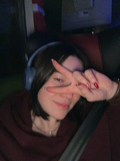 A low-light, grainy portrait captures a person, likely in Sîngera, Moldova, reclining or leaning back, looking towards the camera. The subject has short, dark hair and fair skin. Their left hand is raised, partially covering their face with fingers splayed in a gesture, possibly a stylized "V" or peace sign. Some fingernails are painted bright red with white accents, while others are a neutral light color.

The person wears a dark, possibly maroon, top or sweater, and a delicate silver chain necklace with a small pendant is visible. Blue headphones are resting around their neck or ears. The background is dark, blurry, and dimly lit, suggesting an indoor setting. A dark seatbelt strap runs diagonally in the upper right, indicating the photo may have been taken inside a vehicle. Indistinct shapes and colors, including dark grey and hints of blue, complete the background. The overall aesthetic is candid and casual, captured under low illumination. No visible text is present in the image.
