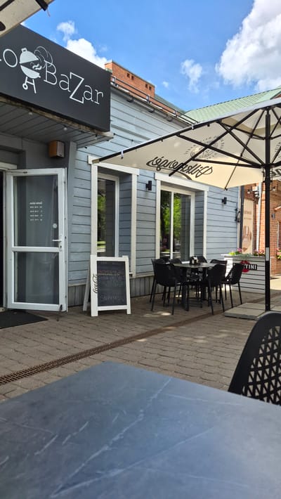 The image shows the outdoor seating area of a restaurant or cafe named "o BaZaR" in Zarasai, Lithuania. The facade of the building is light grey wood paneling. A black sign with white lettering "o BaZaR" featuring a grill graphic is mounted above the entrance. An open glass door reveals a sign with opening hours in Lithuanian. Adjacent to the door is a white A-frame sign with a handwritten menu in Lithuanian.

In the foreground, a dark grey table with a marble-like pattern is visible. Behind it, several black plastic chairs surround a square table under a large white umbrella with black lettering that is partially visible. The umbrella is decorated with a repeating pattern. The ground is paved with light brown cobblestones.

To the right of the seating area, a white counter with flowers and a "MARTINI" logo is present. In the background, a brick building with a green roof is visible, along with trees and a blue sky with white clouds. The overall scene depicts a sunny day in a relaxed outdoor dining setting. There are no people visible in the image.